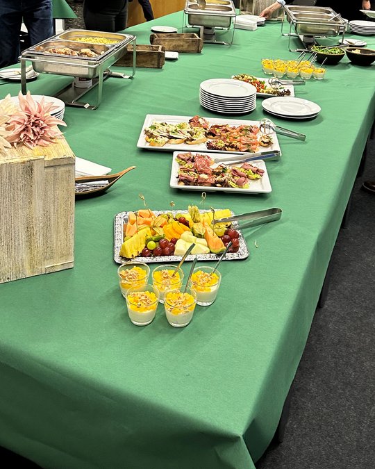 A buffet table covered with a green tablecloth displays various dishes. There are platters of colorful fruit, savory appetizers, small cups with dessert, and stacks of white plates. In the background, other people can be seen serving themselves.