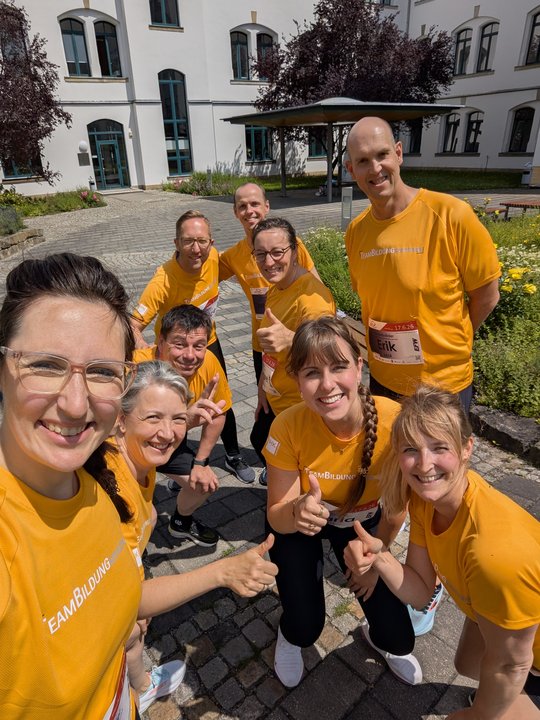 A group of nine people dressed in matching yellow t-shirts, some with event tags, pose together outdoors. They smile and give thumbs up, standing in a courtyard with greenery and a building in the background. The atmosphere is cheerful and energetic.