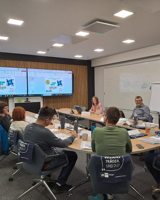 A group of seven individuals are seated around a conference table in a modern meeting room. One person, a woman with long hair, speaks while two large screens display visual content. Participants are engaged in discussion, with notebooks and reusable water bottles visible on the table.