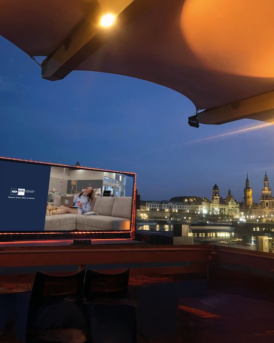 Ein Fernseher zeigt eine Frau, die entspannt auf einer Couch sitzt und am Laptop arbeitet. Im Hintergrund ist eine beleuchtete Stadtlandschaft bei Nacht zu sehen. Die Szene vermittelt eine ruhige, moderne Atmosphäre.
