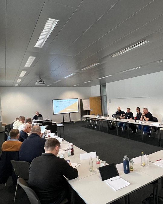A group of people are seated around a conference table, engaged in discussion. A presentation screen displays information in the background. Participants appear to be taking notes or using devices, indicating an interactive meeting environment. The room is well-lit and modern.