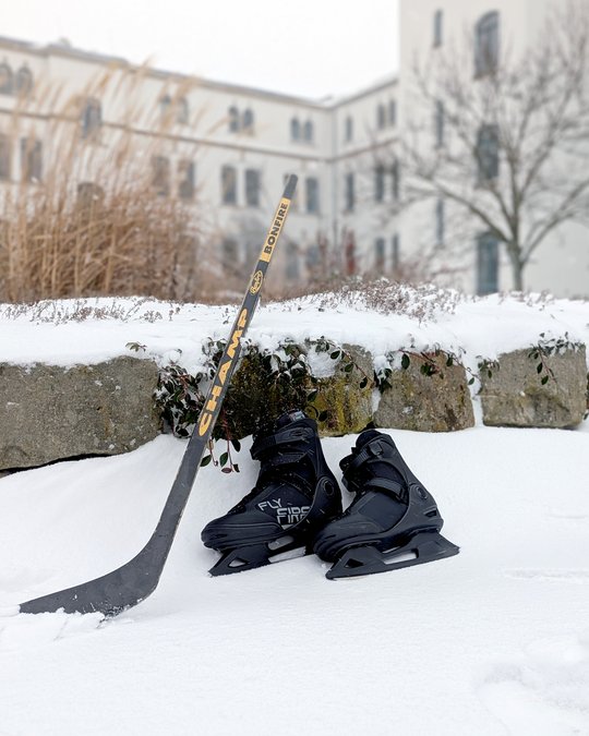 In a snowy landscape, a pair of black hockey skates sits on the ground next to a hockey stick. The scene conveys a sense of winter sports and outdoor activity. The background features blurred buildings and some winter vegetation.