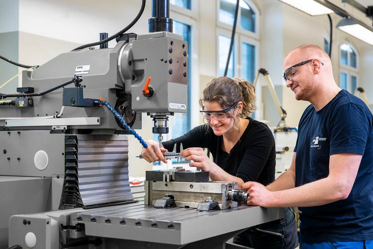 Two individuals are working together in a workshop, using a milling machine. One person, a woman, is adjusting a piece of material, while the other, a man, observes. Both are wearing safety glasses and appear engaged in their task, with bright, well-lit surroundings.