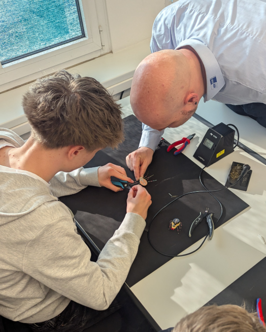 A male instructor leans over a desk, guiding a young male student as they work together on a small electronic project. Tools and components are laid out on a black surface, while a soldering iron and other equipment are visible nearby. Natural light streams in from a window.