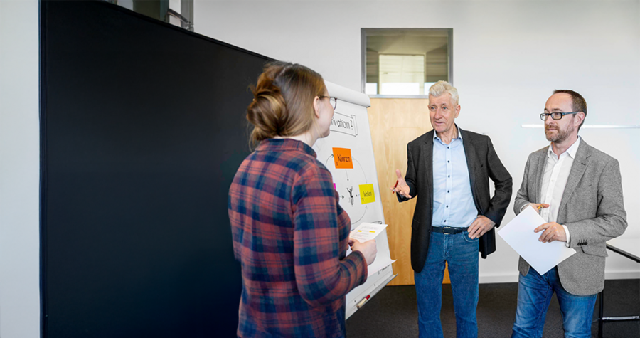 A woman in a plaid shirt stands facing two men in a conference room. One man, with gray hair, gestures while speaking, and the other holds papers. A whiteboard with written content is visible in the background. The scene depicts a collaborative discussion or presentation.