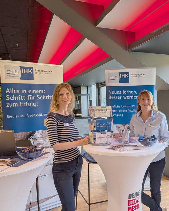 Two women stand behind a table at an event, promoting educational programs. They smile and engage with visitors. The backdrop features banners highlighting the IHK (Chamber of Commerce) with messages about success in training and development. The setting is bright and modern.