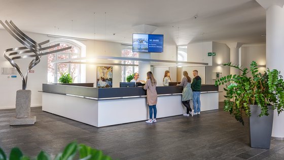 A modern reception area featuring a sleek desk with people interacting with staff. A digital screen displays information above the desk. Decorative plants and a sculptural art piece are present, creating a welcoming atmosphere. Bright windows provide natural light.