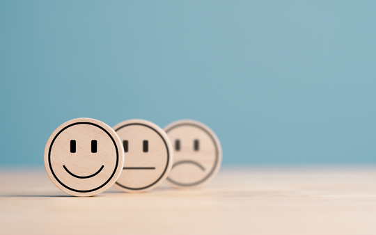 Three wooden tokens with smiley faces are displayed against a light blue background. The first token shows a happy face, the second a neutral face, and the third a sad face, representing a range of emotions on a simple, clean surface.