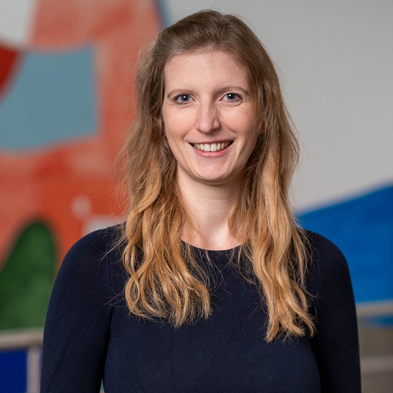 A woman with long, wavy hair smiles confidently at the camera. She is wearing a dark top and stands against a colorful abstract background. Her expression is warm and inviting, suggesting a friendly demeanor.