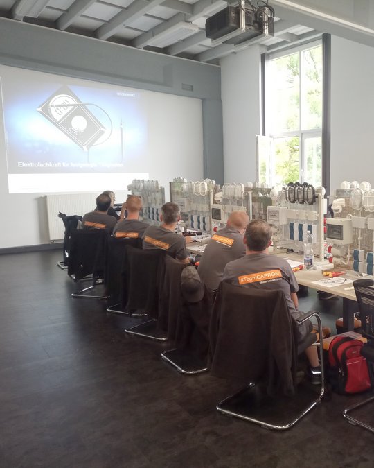 In einem modernen Schulungsraum sitzen mehrere Personen in grauen T-Shirts mit der Aufschrift "#TeamCAPRON" an einem Tisch. An der Wand ist eine Präsentation zum Thema Elektrofachkraft zu sehen. Der Raum ist hell, mit großen Fenstern und einer technischen Ausstattung auf dem Tisch.