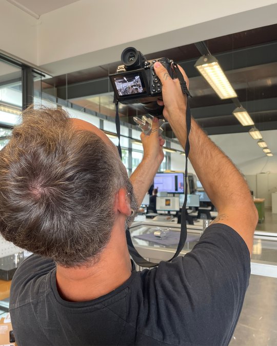 Ein Mann, der von hinten zu sehen ist, hält eine Kamera in der Hand, während er etwas fotografiert. Im Hintergrund sind Arbeitsplätze und ein Computerbildschirm zu erkennen. Der Mann trägt ein schwarzes T-Shirt und scheint sich auf das Fotografieren zu konzentrieren.