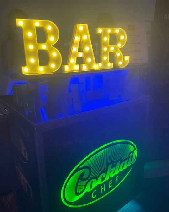 Leuchtende Buchstaben bilden das Wort "BAR" auf einer Cocktailtheke, die im Hintergrund blau beleuchtet ist. Darunter ist das Logo "Cocktail Chef" in grünem Licht sichtbar. Der Fokus liegt auf der einladenden Barumgebung.
