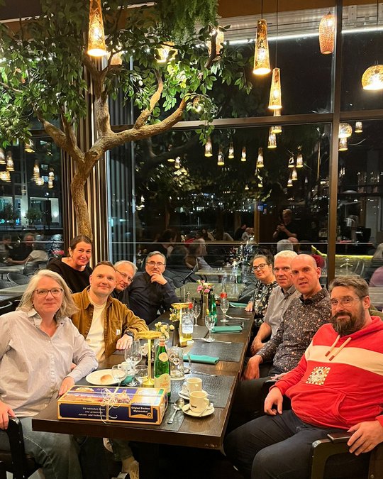 Eine Gruppe von neun Personen sitzt an einem schön gedeckten Tisch in einem Restaurant. Auf dem Tisch sind Getränke und Blumenvase sichtbar. Im Hintergrund sind große Fenster und eine Begrünung zu sehen. Die Atmosphäre ist gemütlich und einladend.