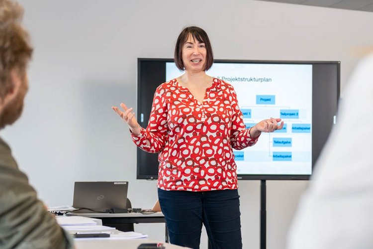 A woman in a red and white polka dot shirt is standing in front of a presentation screen, engaging an audience seated at a table. She has her arms raised as she speaks. The setting appears to be a modern classroom or meeting room.