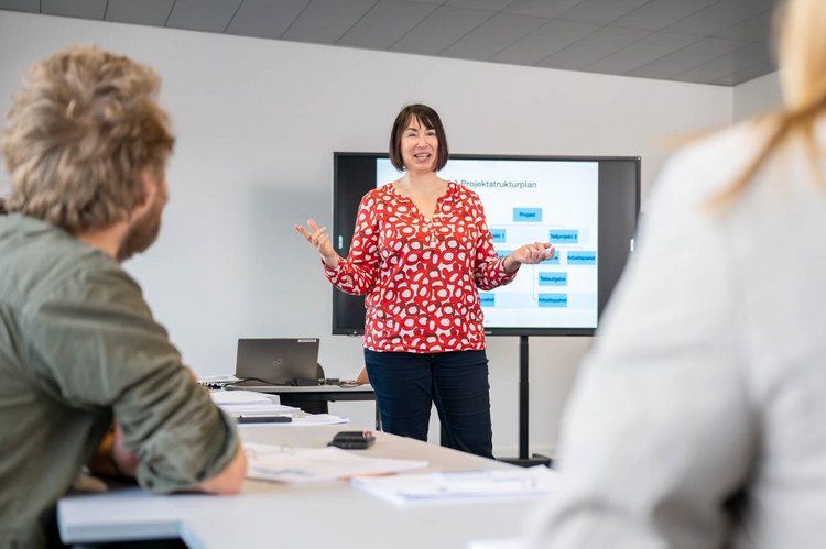 A woman in a red and white polka dot shirt is standing in front of a presentation screen, engaging an audience seated at a table. She has her arms raised as she speaks. The setting appears to be a modern classroom or meeting room.