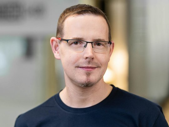 Porträt eines Mannes mit kurzen, hellen Haaren und einer leichten Gesichtsbehaarung. Er trägt ein dunkelblaues T-Shirt mit einem Logo. Der Hintergrund ist unscharf, wobei eine moderne Umgebung angedeutet wird. Der Mann lächelt leicht und sieht freundlich aus.