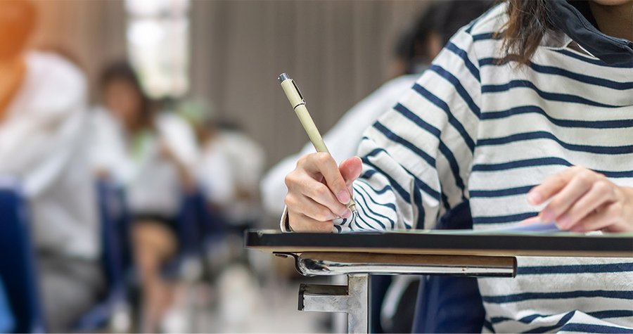 A close-up of a person wearing a striped shirt, holding a pen and writing on a piece of paper at a desk. In the background, other students are seated, suggesting a classroom or examination setting. The focus is on the act of writing.