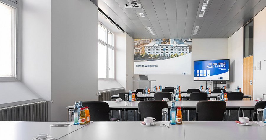 Ein moderner Konferenzraum mit mehreren Tischen, die für ein Treffen vorbereitet sind. Auf jedem Tisch stehen Wasserflaschen und Tassen. An der Wand befindet sich ein Begrüßungsschild und ein Bildschirm, der Informationen anzeigt. Viel Tageslicht fällt durch die Fenster.