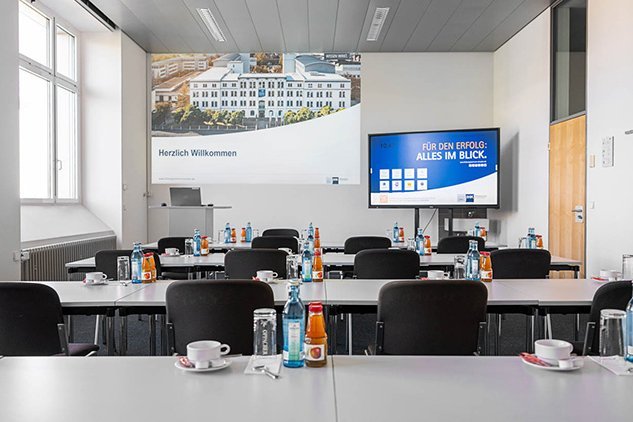 Ein moderner Konferenzraum mit mehreren Tischen, die für ein Treffen vorbereitet sind. Auf jedem Tisch stehen Wasserflaschen und Tassen. An der Wand befindet sich ein Begrüßungsschild und ein Bildschirm, der Informationen anzeigt. Viel Tageslicht fällt durch die Fenster.