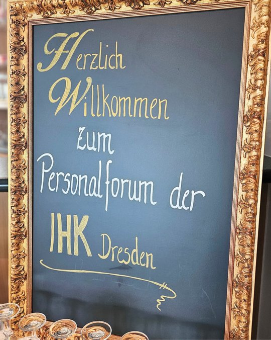 Eine elegante Tafel mit dem Text: "Herzlich Willkommen zum Personalforum der IHK Dresden". Der Text ist in goldener Schrift auf dunklem Hintergrund geschrieben, umgeben von einem verzierten Rahmen.