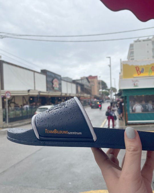 A person holds a dark blue slide sandal with the text "TEAMBILDUNGszentrum" displayed on it. In the background, a street scene features buildings, people walking, and cloudy skies indicating rain. The setting suggests a casual, urban environment.