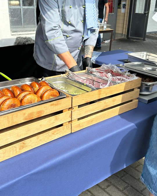 Ein Koch in grauer Uniform bereitet Essen an einem Tisch vor. Vor ihm befinden sich mehrere Edelstahlbehälter mit verschiedenen Speisen, darunter frische Brötchen. Der Tisch ist mit einer blauen Tischdecke bedeckt. Der Koch trägt Handschuhe und hat ein Tuch um den Hals.