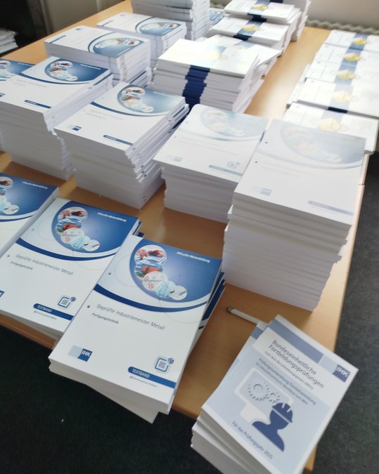 Auf dem Tisch liegen Stapel von Dokumenten mit verschiedenen Titeln, darunter „Geprüfte Industriemeister Metall“ und „Bundeseinheitliche Fortbildungsprüfungen“. Die Unterlagen sind ordentlich sortiert und einige sind mit blauen Bändern gebunden. Die Auflage scheint für Schulungen oder Prüfungen im Bereich Metall zu sein.