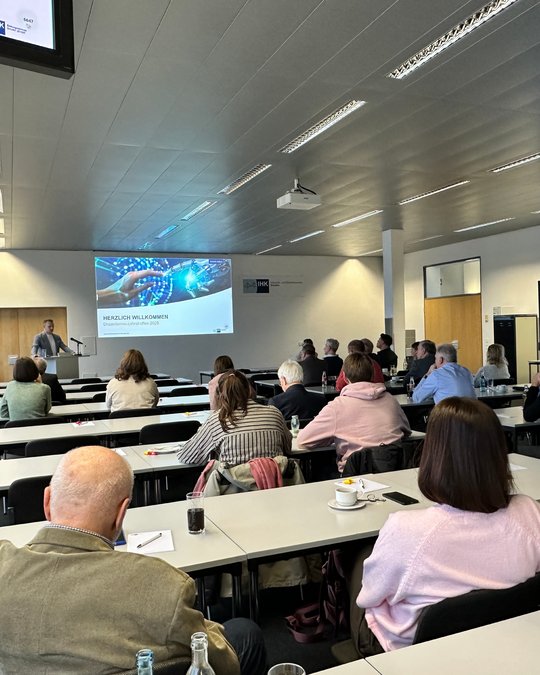 A presenter stands at the front of a conference room, addressing an audience seated at tables. A large screen displays "WELCOME" alongside a colorful graphic. Attendees, mostly adults, are engaged with the presentation, creating an atmosphere of collaboration and learning.