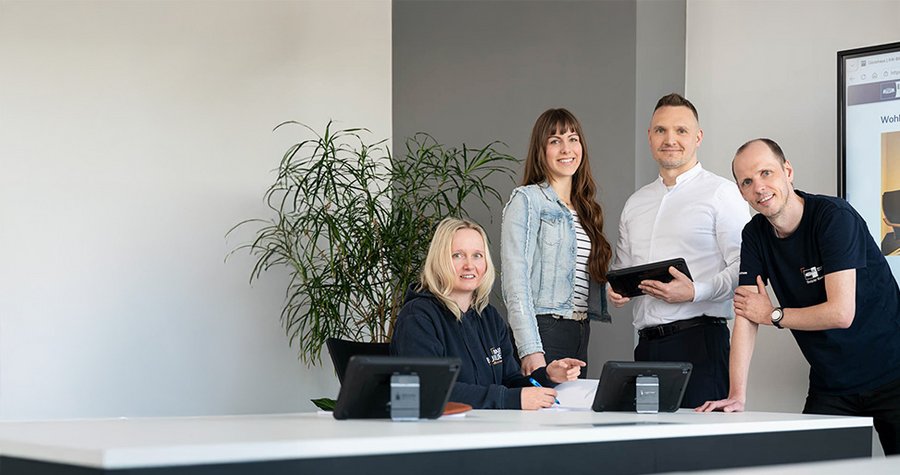 A group of four professionals stands around a modern reception desk. One person is seated, taking notes, while the others engage with electronic devices. The background features a plant and a screen displaying information. The atmosphere is collaborative and welcoming.