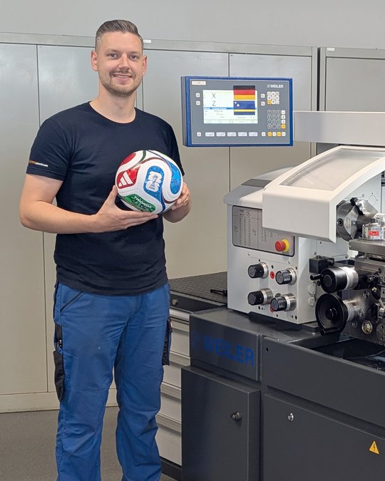 A man stands next to a printing machine, holding a colorful soccer ball. He is wearing a black shirt and blue pants, with a friendly smile. The machine features a digital control panel with various buttons and displays, indicating a manufacturing or printing setting.