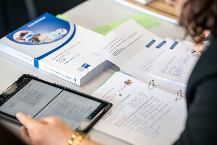 A person's hands are holding a tablet displaying text, while a textbook and organized notes are visible on the table. The scene suggests a study or research setting, focusing on learning materials and digital resources.