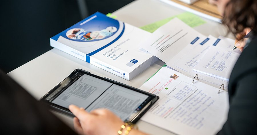 A person studying at a table with a tablet displaying digital text and a textbook nearby. There are also handwritten notes and documents spread out, suggesting a focus on learning and organization.
