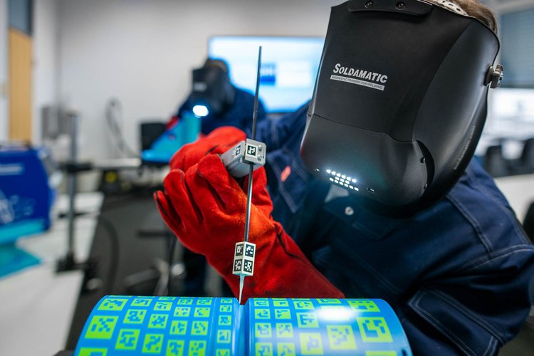 A person wearing a welding helmet and red gloves is holding a metal piece with a measurement tool, focusing on precision welding. In the foreground, there are vibrant blue and green materials, indicating a workspace environment for welding or metal fabrication.