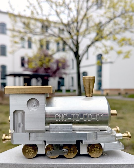 A small, metallic model of a steam locomotive is displayed in the foreground. The train features a shiny silver body with golden accents, including a smokestack. It is set against a blurred background of a building and trees, suggesting an outdoor scene.
