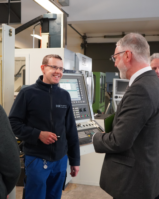 A technician in a blue sweater engages in conversation with a man in a suit, both smiling. The background features industrial machinery, suggesting a workshop or manufacturing setting. The interaction appears friendly and professional, highlighting an exchange of ideas or information.