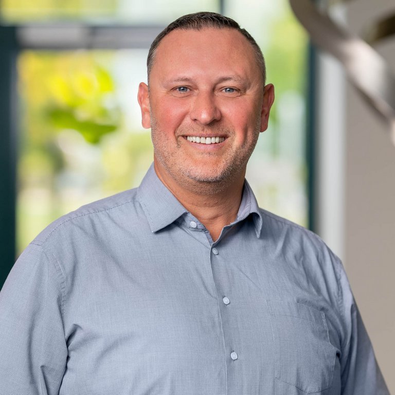A man with short, neatly styled hair smiles confidently at the camera. He is wearing a light blue collared shirt. The background features soft blurred greenery and indoor decor, creating a warm and inviting atmosphere.