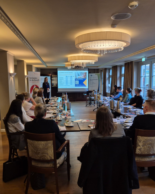 A professional woman stands at the front of a well-lit conference room, presenting information on a screen. Attendees are seated at a long table, engaged and listening. The room has a modern decor with large windows and decorative lighting.