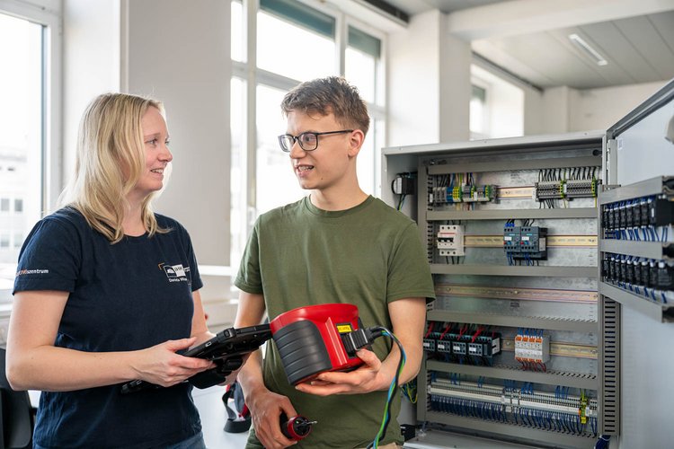 Two individuals are engaged in discussion in a room filled with natural light. One person, a woman with long hair, holds a tablet, while a man with glasses holds a red device. Behind them is a control panel featuring various electrical components.