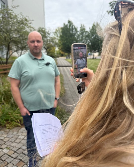 A man stands outdoors, looking towards the camera, while a person with long hair faces him, holding a smartphone for recording. The background features some greenery and a pathway. The scene appears to capture an interview or presentation.