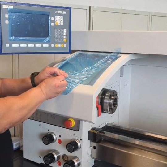 Eine Person entfernt eine Schutzfolie von der Bedienoberfläche einer Maschine. Die Maschine verfügt über einen Bildschirm und mehrere Knöpfe auf der Vorderseite. Im Hintergrund sind weitere Maschinen sichtbar.