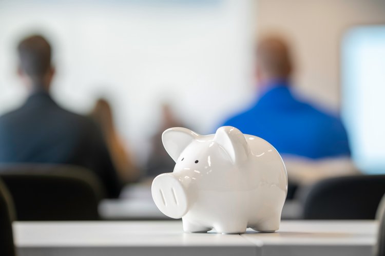 A white piggy bank sits on a table in the foreground, symbolizing savings and financial planning. In the blurred background, a group of people can be seen attending a presentation or meeting.