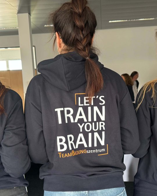 Auf einem Rückensweatshirt steht der Slogan: „LET'S TRAIN YOUR BRAIN“ in großen, auffälligen Buchstaben. Darunter befindet sich der Schriftzug „TeamBildungszentrum“. Die Personen sind von hinten zu sehen, wobei sich mehrere Menschen in der Nähe befinden.