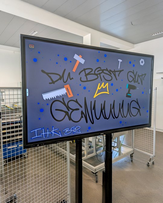 A digital screen displays colorful graffiti-style text that reads "DU BEST GUT GENUUUUUG." Various graphic elements, such as tools and a crown, are incorporated into the design. The background includes blue splashes. The screen is mounted on a black stand, situated in a light-colored room.