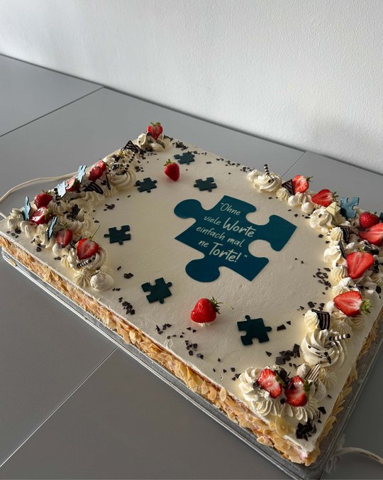 Eine rechteckige Torte mit einer hellen Sahnefüllung, dekoriert mit Erdbeeren, Schokoladenstückchen und Sahnetupfen. In der Mitte steht der Text: "Ohne viele Worte einfach mal ne Torte!" auf einem Puzzlestück.
