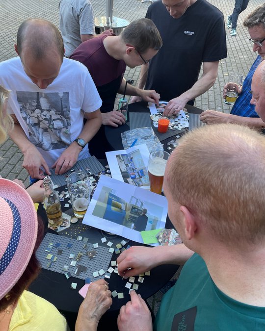 Eine Gruppe von Menschen sitzt an einem Tisch und arbeitet gemeinsam an einem Puzzlespiel. Vor ihnen liegen verschiedene Puzzlestücke und Bilder, die als Referenz dienen. Einige Personen trinken Getränke. Der Fokus liegt auf dem gemeinsamen Spiel und der Interaktion zwischen den Teilnehmern.