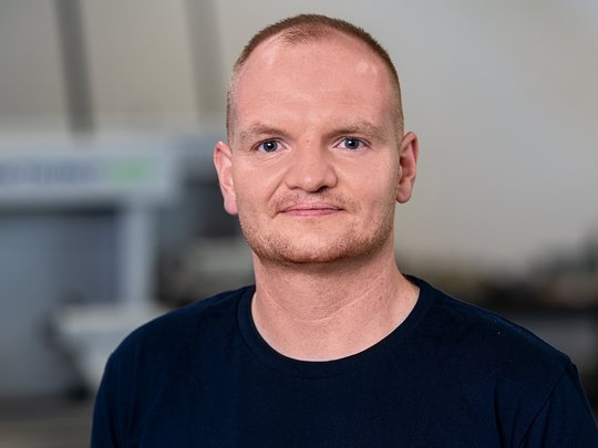 A man with short, light-brown hair is wearing a black T-shirt with a logo and his name, "Martin Deutsch." He is looking directly at the camera, smiling, in a workshop setting with blurred machinery in the background.