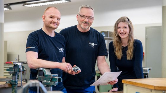 Three people stand together in a workshop, smiling and engaged in discussion. They hold tools and a document, surrounded by workbenches and equipment. The scene suggests a collaborative atmosphere focused on craftsmanship or engineering.
