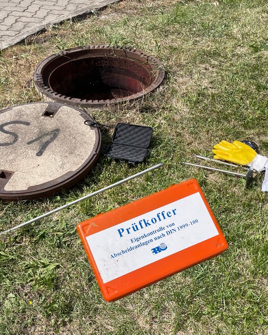 Das Bild zeigt einen geöffneten Kanaldeckel mit einem überprüfenden Koffer auf dem Boden, der mit „Prüfkoffer“ und Informationen zur Eigenkontrolle von Abscheideanlagen gekennzeichnet ist. Außerdem sind Teststäbe und ein Paar gelbe Handschuhe zu sehen. Grass und ein Gehweg umgeben die Szene.