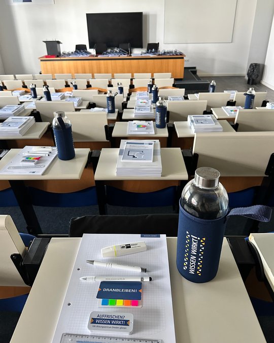 Ein Seminarraum mit mehreren Sitzplätzen, jeder ausgestattet mit einem Notizblock, Stiften und einer Wasserflasche. Auf den Tischen liegen Informationsmaterialien und farbige Markierungen. Im Hintergrund steht ein großer Bildschirm.