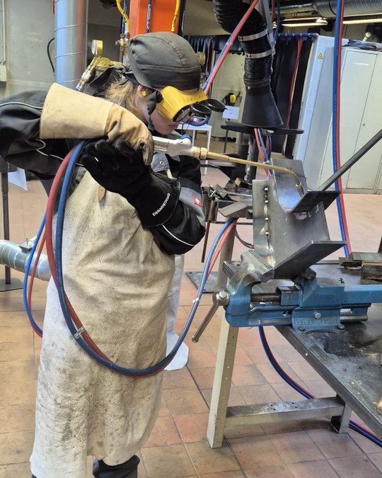 A person in a protective welding outfit is using a welding torch to work on a metal piece fixed on a workbench. The environment appears to be a workshop, with equipment and tools visible in the background.